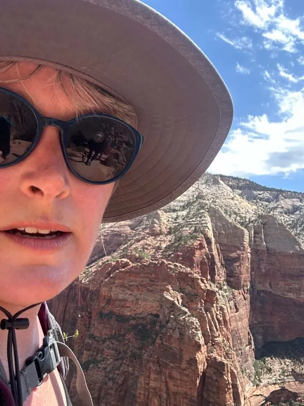 A woman wearing a hat and sunglasses poses at the top of a mountain at Angels Landing in Zion National Park.