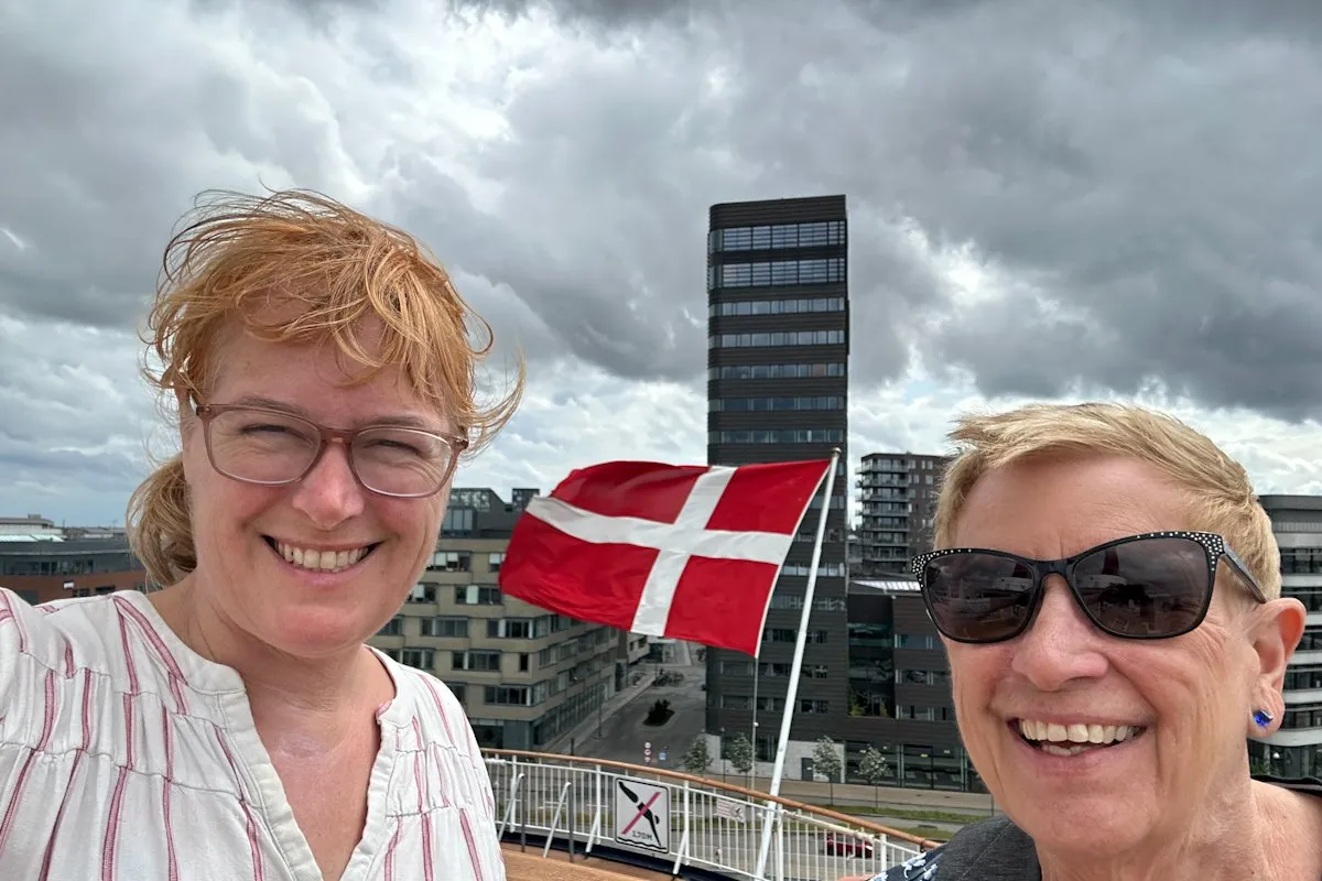 two women smile on a ship with the Danish flag rippling in the wind and Copenhagen's port behind that