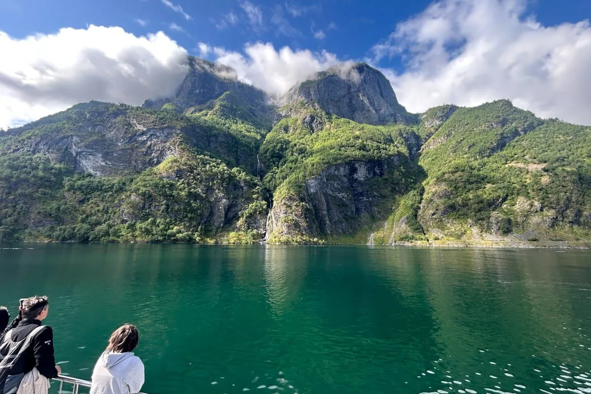 rocky and green mountains with greenish water in the foreground and several people viewing the scenery from a boat